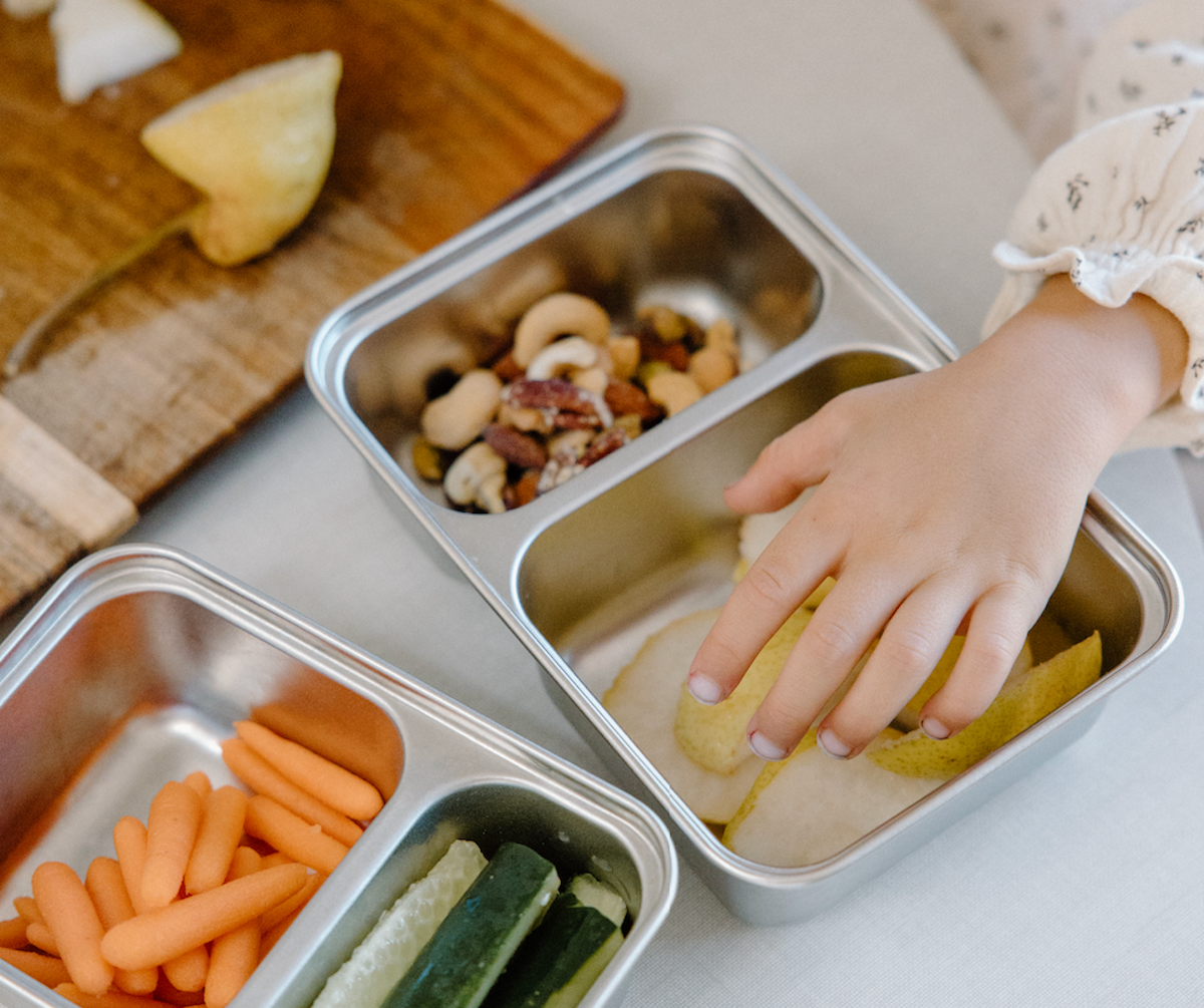 A child’s hands reaching for pears in a stainless steel kids lunchbox.