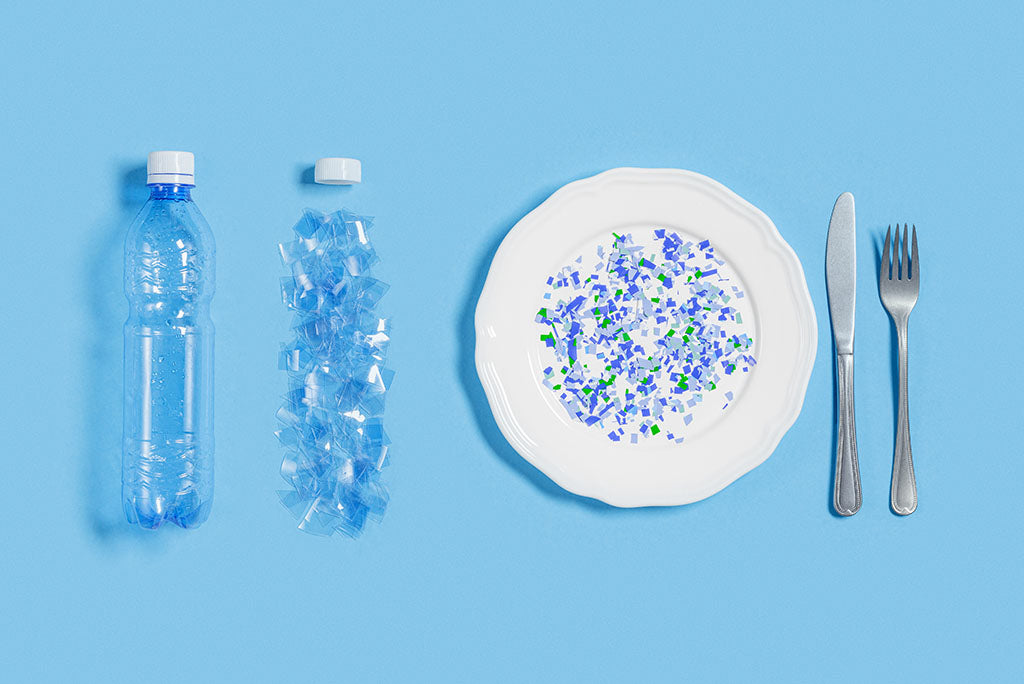 Two plastic water bottles with one in pieces and microplastics on a plate next to a knife and fork.